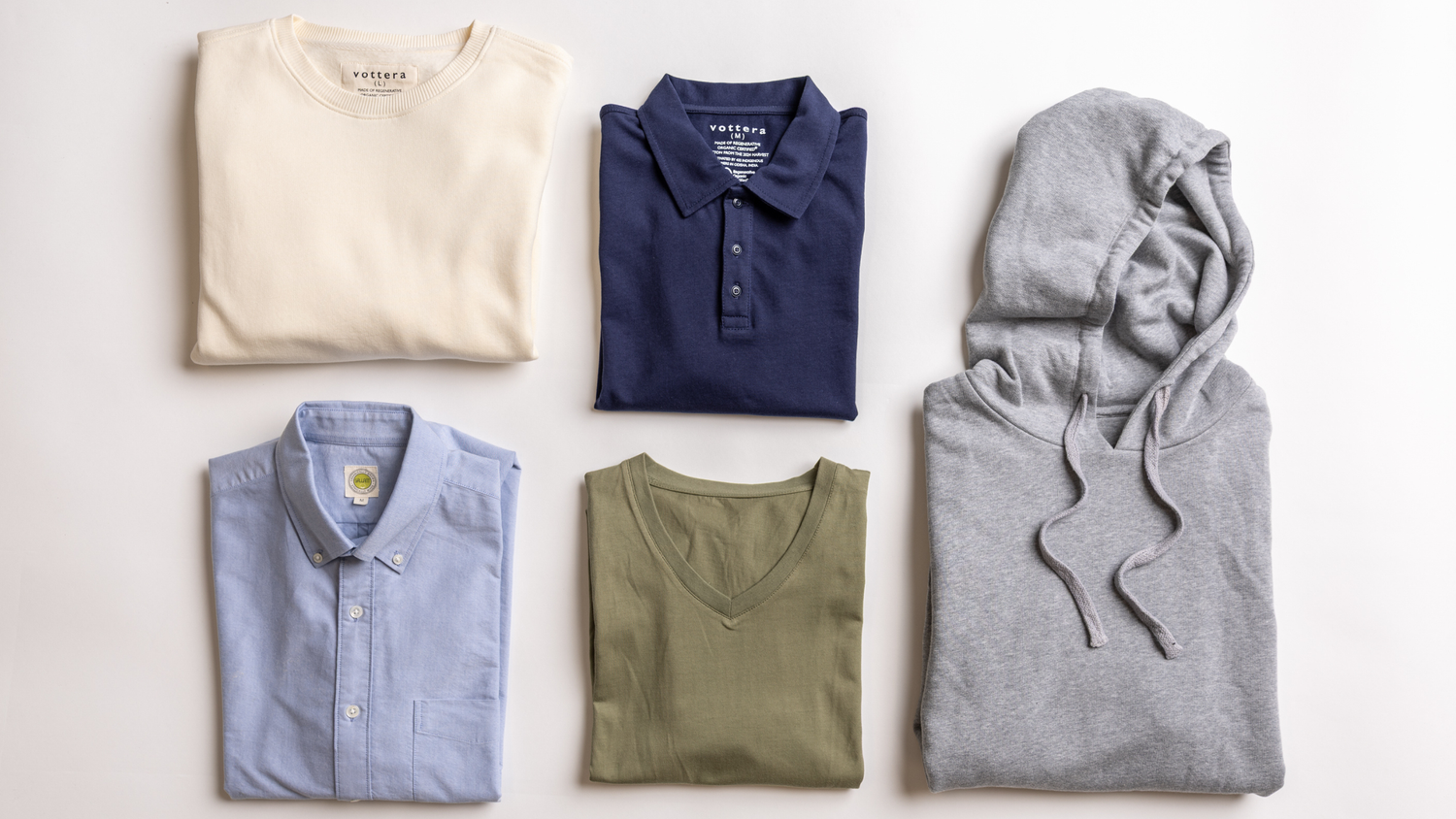 An assortment of slow fashion essentials, including Vottera’s natural color sweatshirt, navy polo, and light blue Oxford shirt made from organic cotton